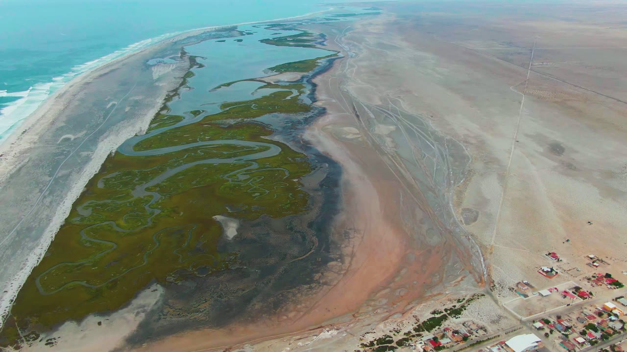 Aerial view over Punta Abreojos, Baja California Sur, with green wetland channels, turquoise ocean at left, pale sand flats at right, distant red-roof buildings, and no visible people.