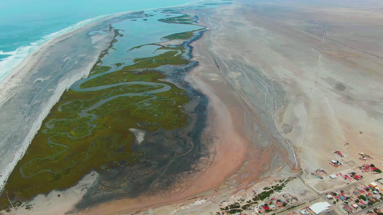 Wide drone view of estuary channels, beach, and small coastal settlement.