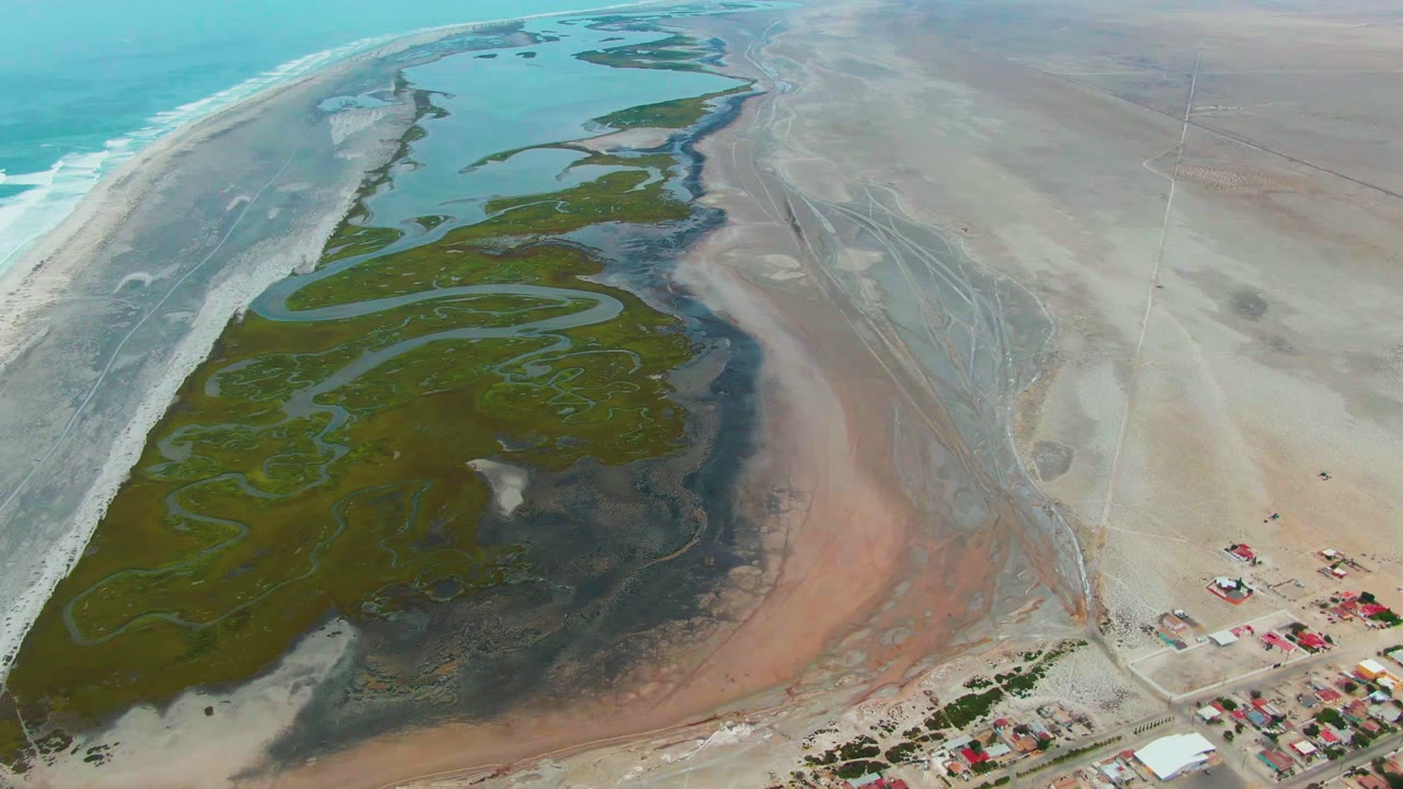 Punta Abreojos aerial frame with the green wetland mass stretching diagonally through the center, darker wet ground around it, pale dry flats on the right, ocean water on the left, and no visible people.