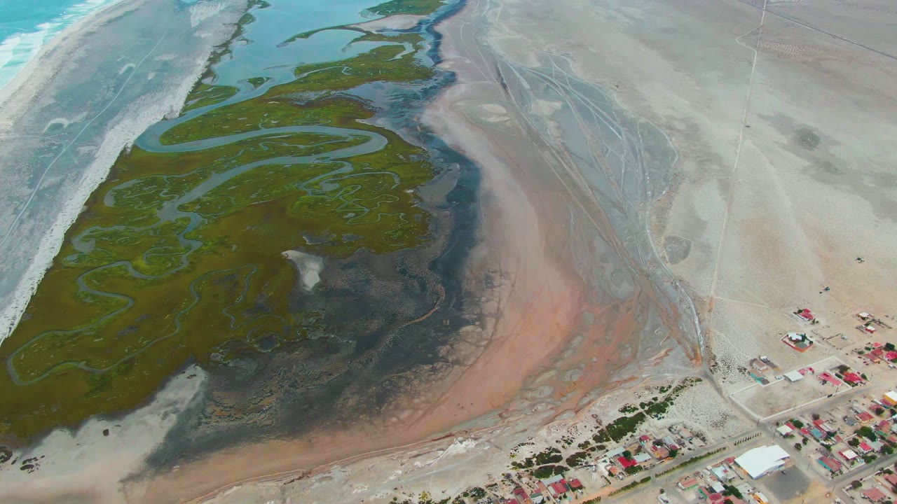 Aerial frame over Punta Abreojos tidal flats showing green algae, winding shallow water, pink-brown sediment, pale sand, edge buildings, and no visible people at the start of the clip.