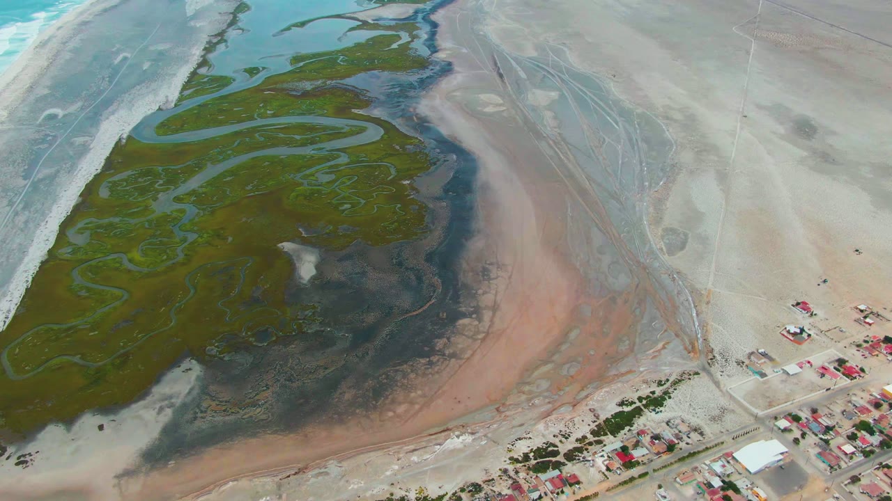 Overhead tidal flat with green algae, dark wet mud, pale sand, and small buildings near the shore.