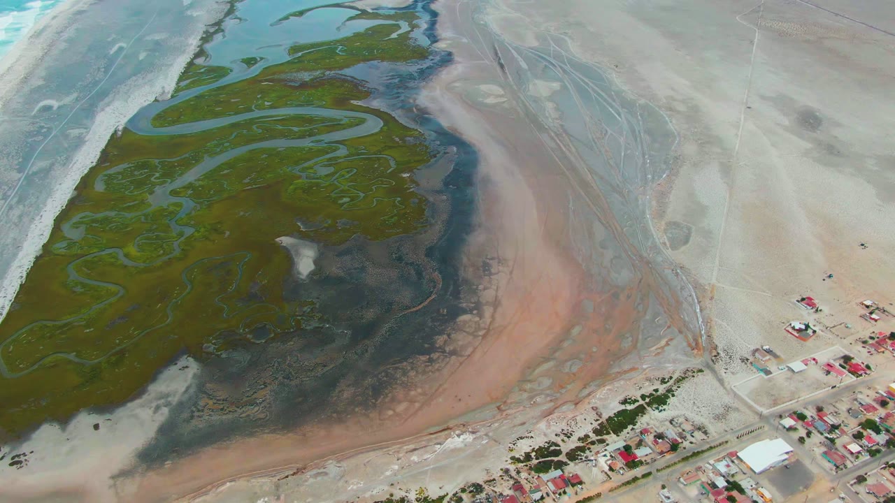 At 0:02, the Punta Abreojos aerial frame shows braided water channels through green algae, a pink sediment band along the exposed flat, small red-roofed buildings at lower right, and no visible people.
