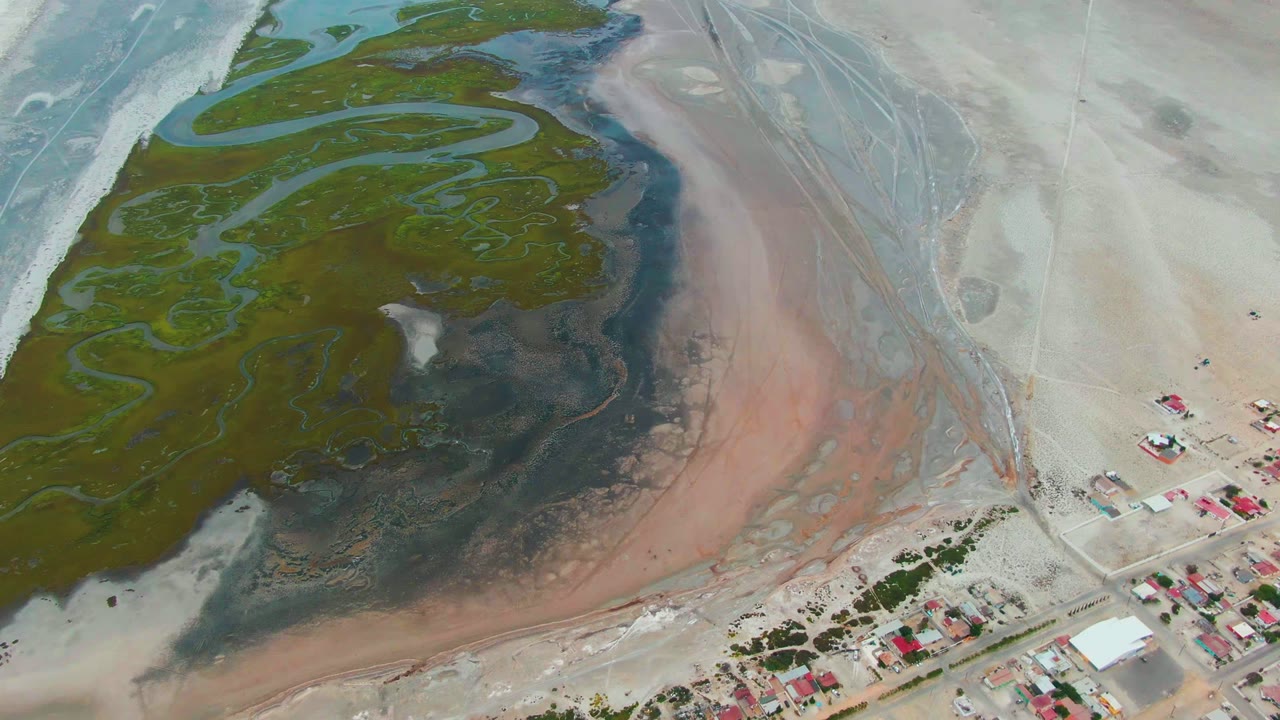 Opening aerial frame over Punta Abreojos, Baja California Sur, showing green lagoon channels, dark shallows, pink water bands, pale sand, a road, red-roofed buildings, and no visible people.