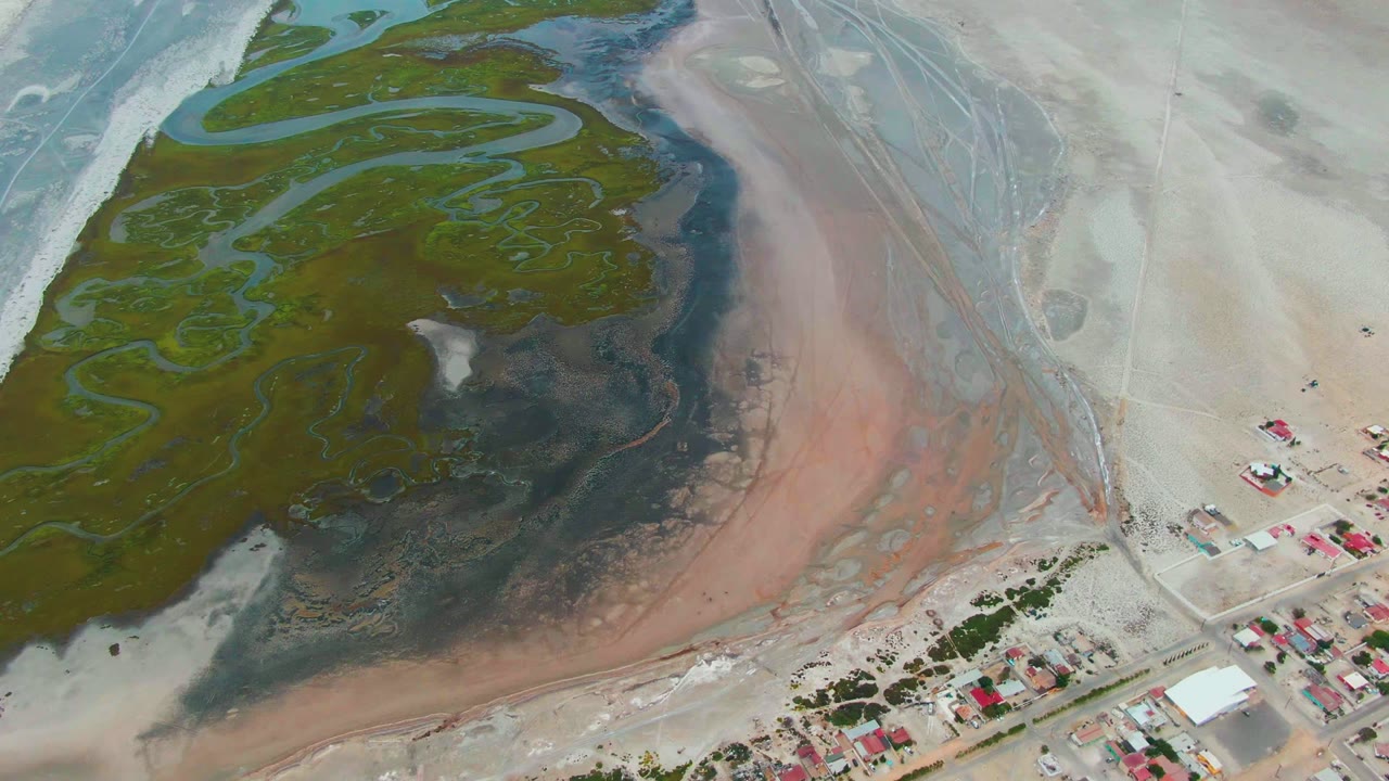 Drone view of colored lagoon water, sandy shore, road, and small buildings with red roofs.