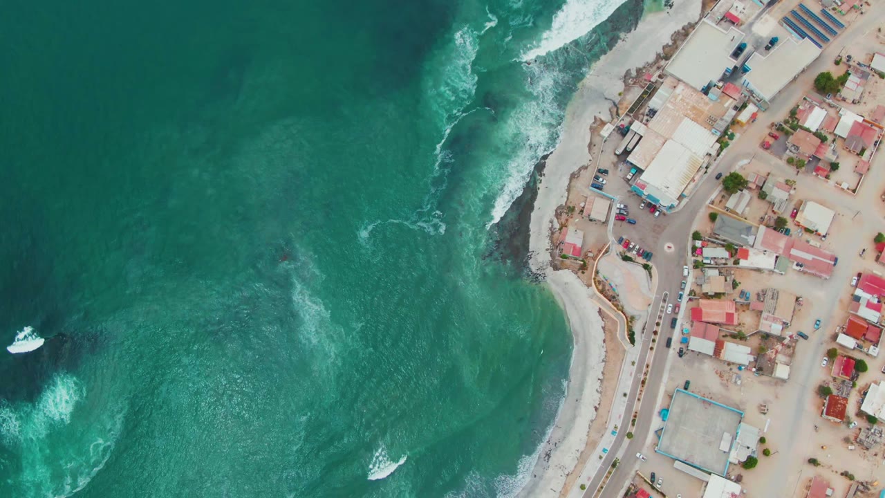 Top-down aerial frame over Punta Abreojos, Baja California Sur, with turquoise ocean on the left, white surf on the beach, roads and low buildings at right, and no visible people.