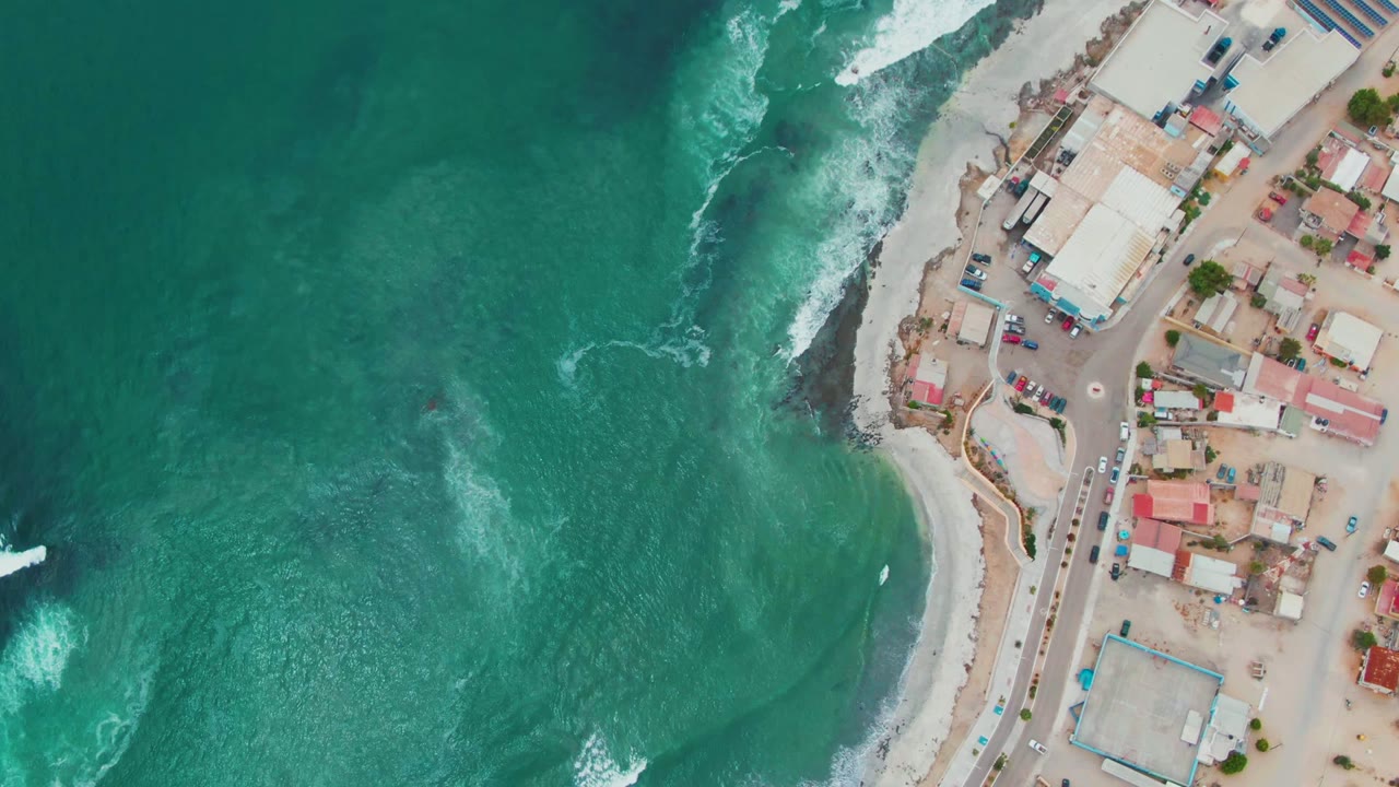 Aerial coastline view with foamy waves curling near a road and clustered roofs.