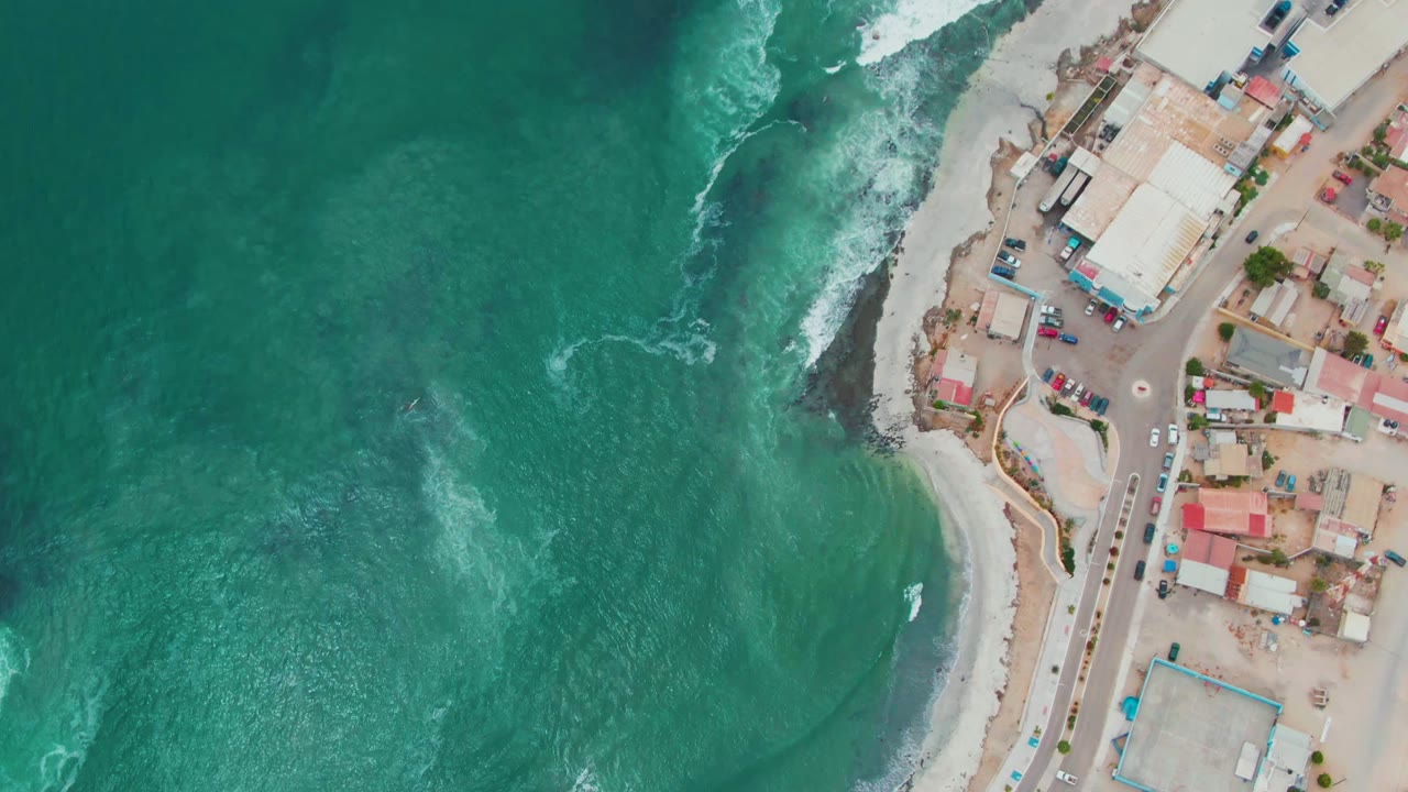 Punta Abreojos aerial frame with textured green-blue water, breaking surf at the beach curve, coastal structures and vehicles along the right edge, and no visible people.
