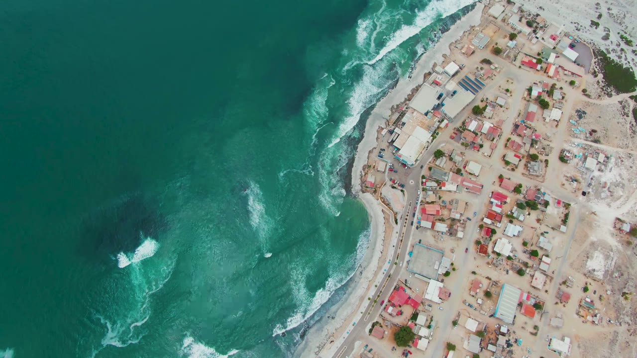 Aerial view over Punta Abreojos, Mexico, with turquoise water on the left, white surf along the shore, a curved coastal road, clustered buildings, and no visible people.