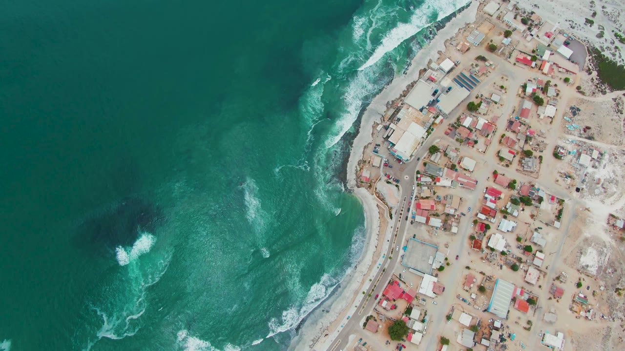 Aerial frame over Punta Abreojos with a broad field of turquoise water, multiple white wave breaks, a rocky shoreline edge, town blocks, and no visible people.