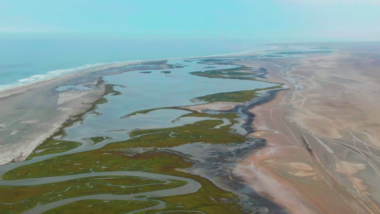 Opening aerial frame over Punta Abreojos shows winding delta channels, green wetland islands, pale sand flats, blue ocean, and no visible people.