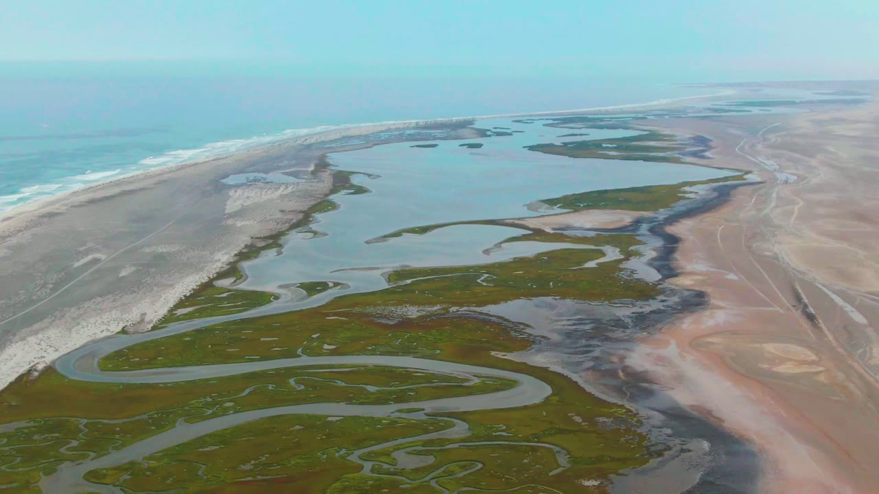 Drone view of meandering water channels crossing marshland near a beach.