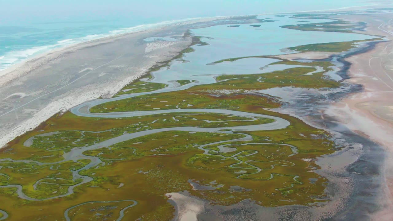 Aerial view in Punta Abreojos with sinuous water channels filling the foreground marsh, pink-gray flats to the right, and a white surf line along the beach, no visible people.
