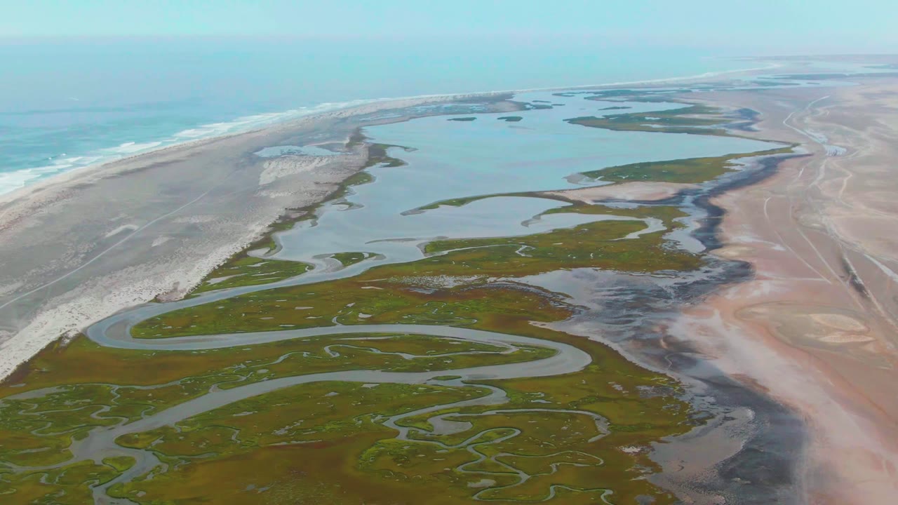 Aerial view over Punta Abreojos, Baja California Sur, with green marsh channels in the foreground, pale sand flats to the right, ocean surf to the left, and no visible people.