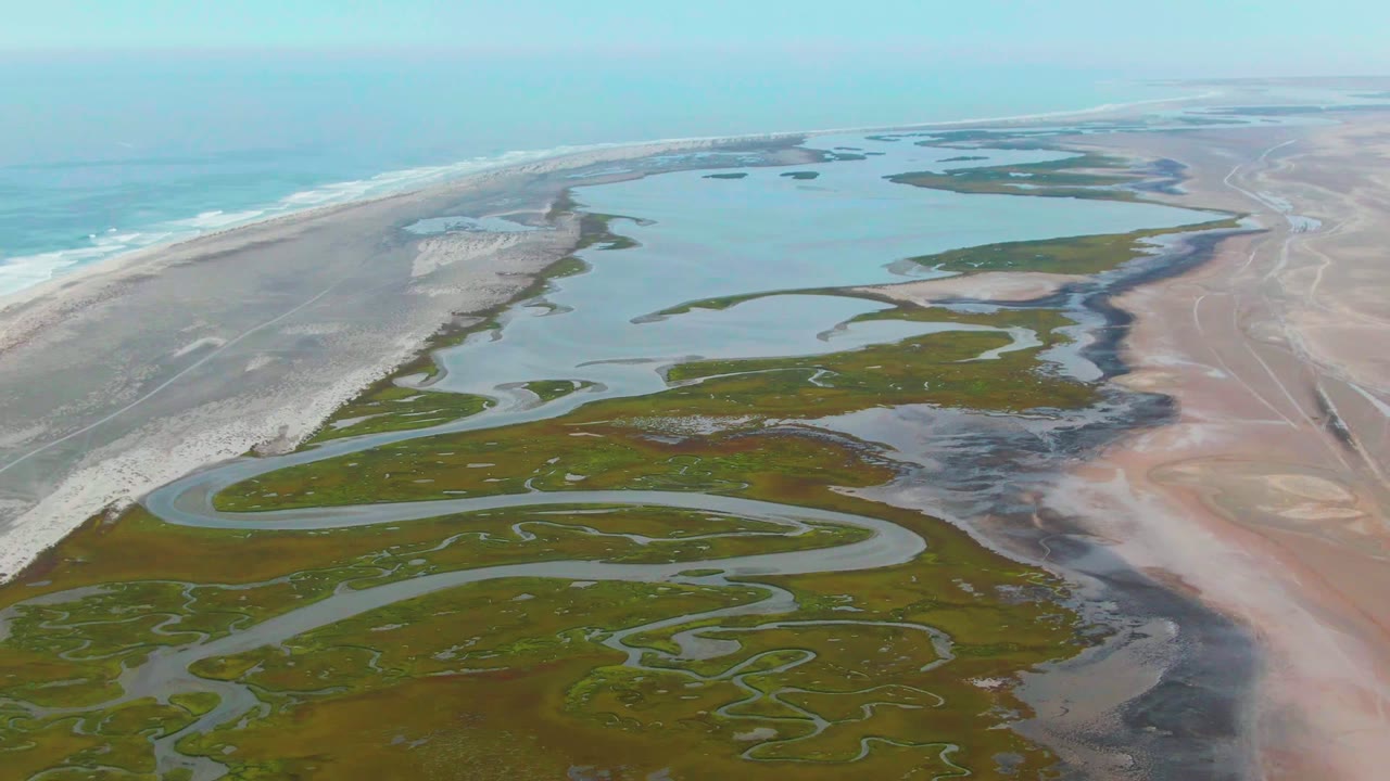 Drone-height view of a coastal wetland delta with ocean surf along the left edge.