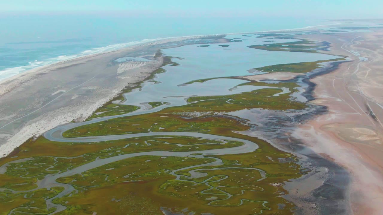 Aerial frame over Punta Abreojos with shallow blue water, scattered marsh islands, pale surf breaking along the outer beach, pink sand flats at right, and no visible people.
