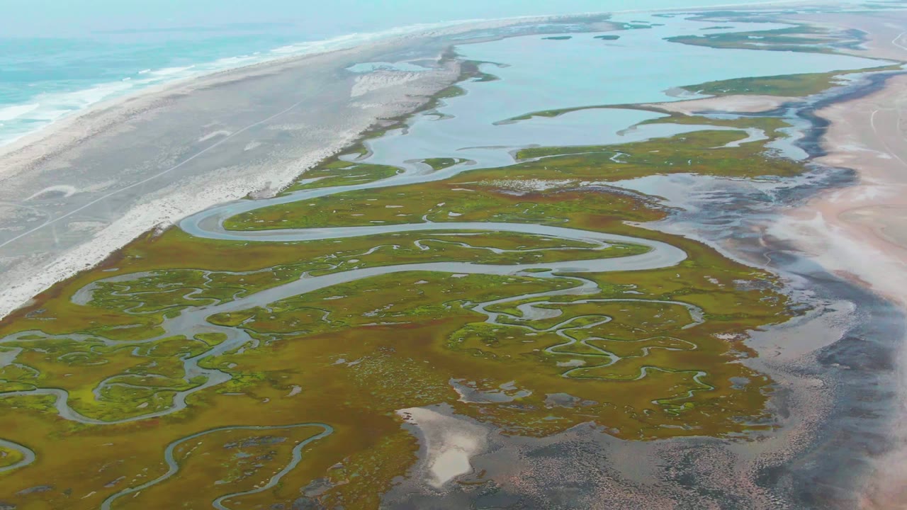 Aerial view over Punta Abreojos marshland with winding blue channels, pale beach, ocean surf at left, shallow water beyond, and no visible people.