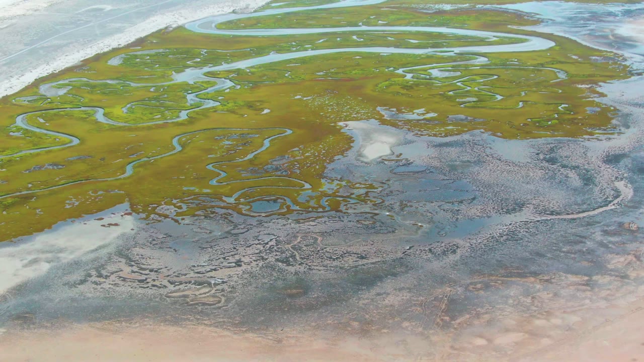 Aerial frame over Punta Abreojos showing looping water channels through green marsh, gray-blue wet mud to the lower right, and tan sand below, with no visible people.
