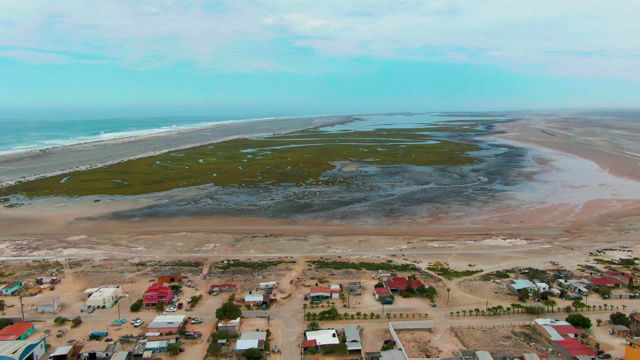 Aerial frame over Punta Abreojos, Baja California Sur, showing houses and sandy roads below tidal wetlands, beach, ocean surf, pale sky, and no visible people.