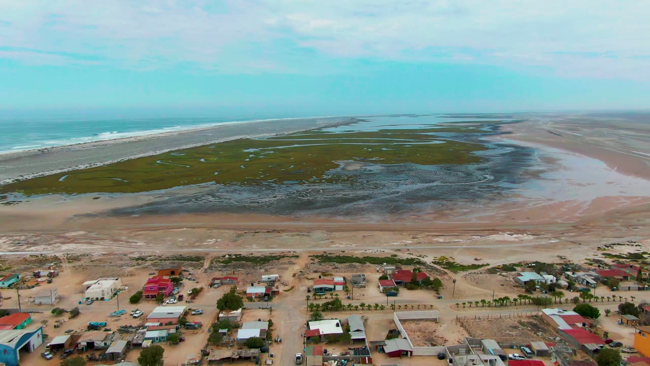 Wide drone frame over Punta Abreojos with colorful roofs in the foreground, patchy tidal flats and wet sand beyond, a long surf line to the left, and no visible people.