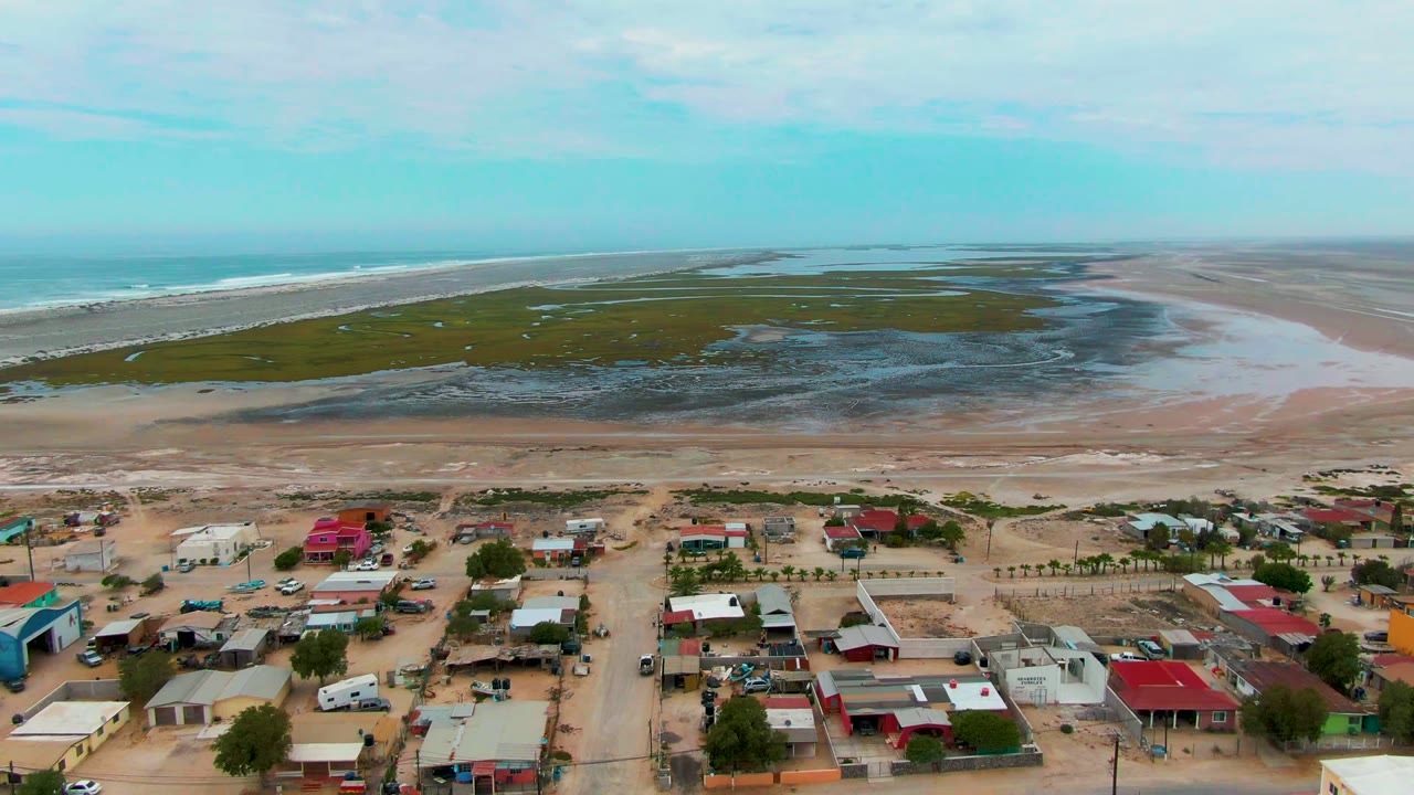Aerial frame over Punta Abreojos, Baja California Sur, showing sandy town streets and low buildings below a beach, green tidal marsh, ocean surf, blue sky, and no visible people.