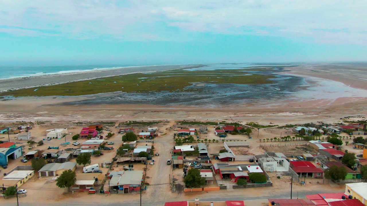 Aerial frame in Punta Abreojos showing red-roofed houses and vehicles at the bottom, a sandy beach strip, tidal marsh pools, ocean waves, and no visible people.