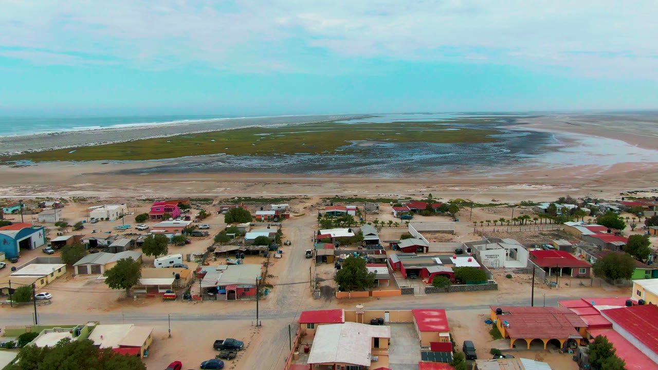 Aerial frame over Punta Abreojos with low houses, red roofs, sandy roads, beach flats, ocean water, partly cloudy sky, and no visible people.