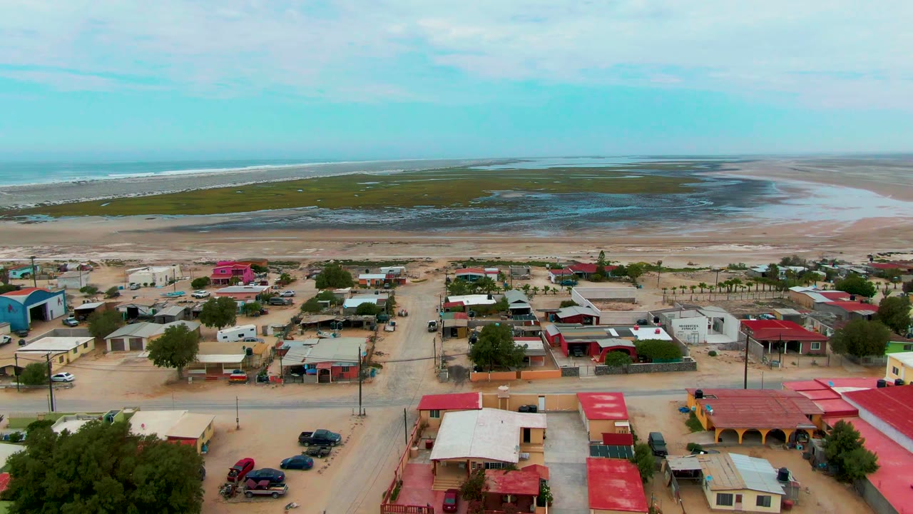 Aerial view of Punta Abreojos with red-roofed and pale buildings in the foreground, parked vehicles, sandy shoreline, ocean horizon, and no visible people.