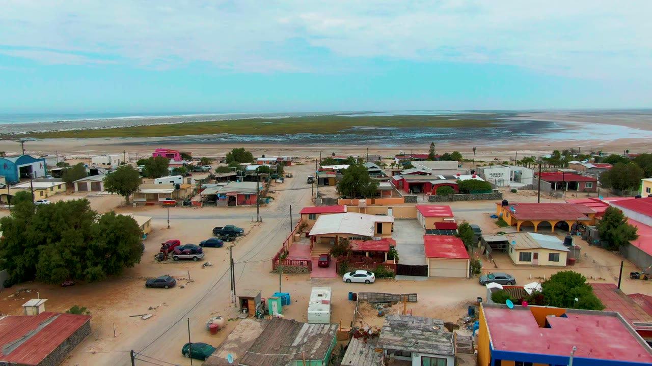 Aerial frame over Punta Abreojos, Baja California Sur, showing sandy streets, red-roofed buildings, parked cars, trees, beach flats, distant ocean, and no clearly visible people.