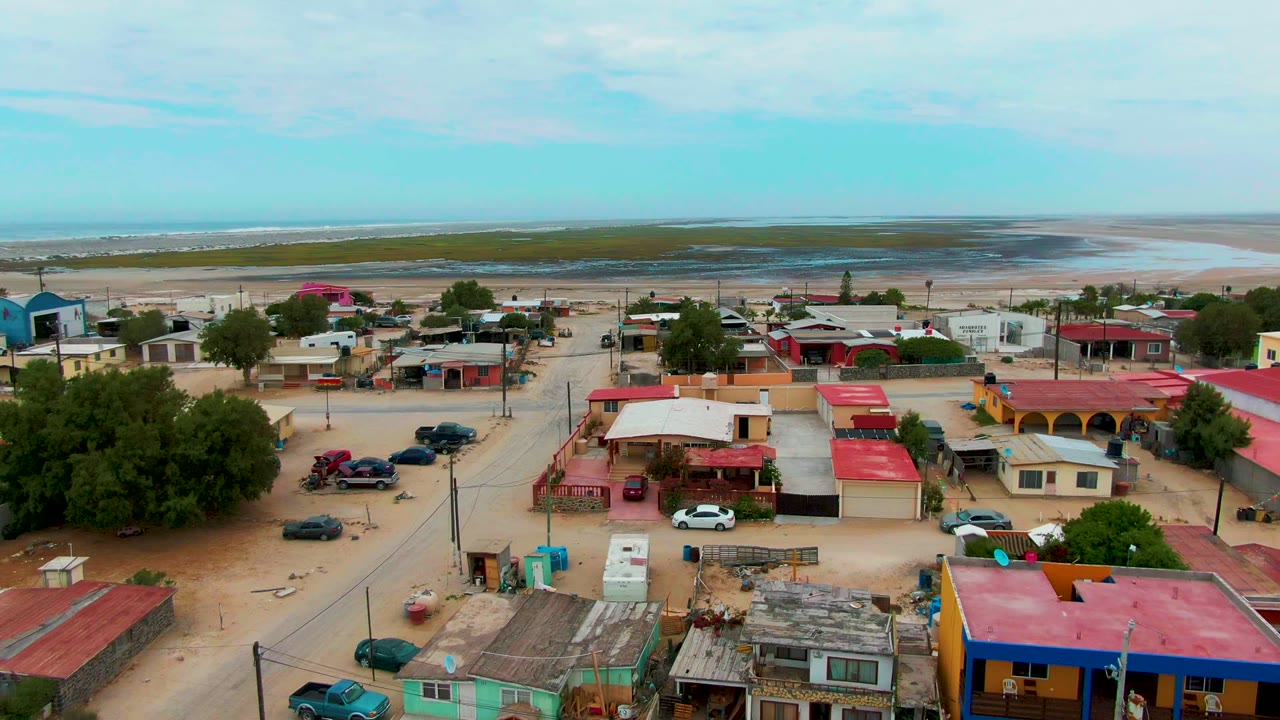 Aerial town view with red roofs, parked vehicles, sandy roads, trees, and ocean in the distance.