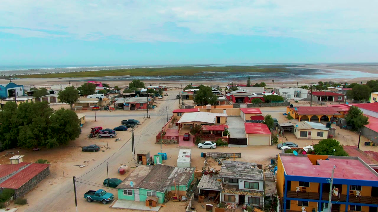 Aerial frame in Punta Abreojos showing colorful low buildings, a white car near the center, sandy roads, trees, pale coastal flats, ocean beyond, and no clearly visible people.