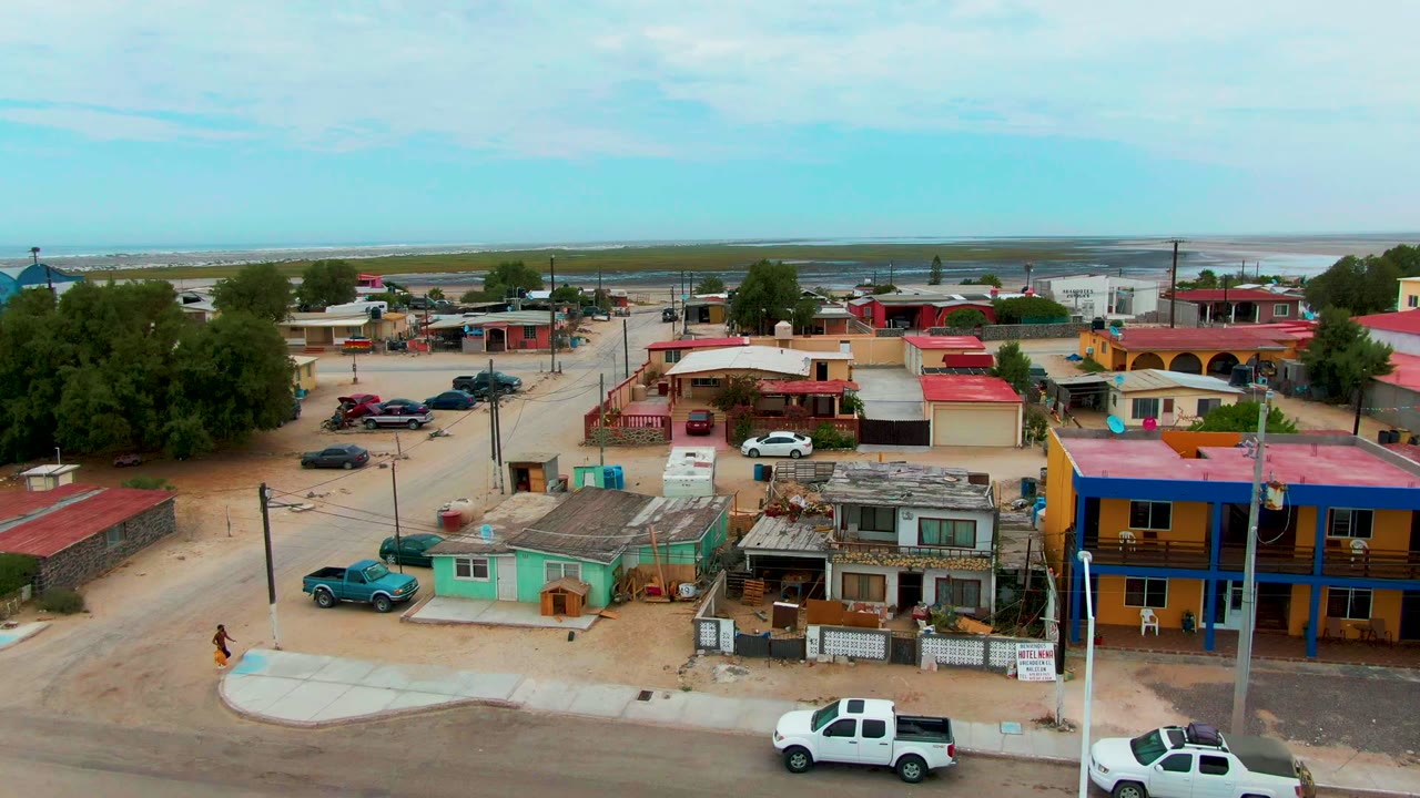 Elevated opening view in Punta Abreojos, Baja California Sur, showing low buildings with red roofs, parked cars, trees, dusty side streets, and a person walking near the lower left.