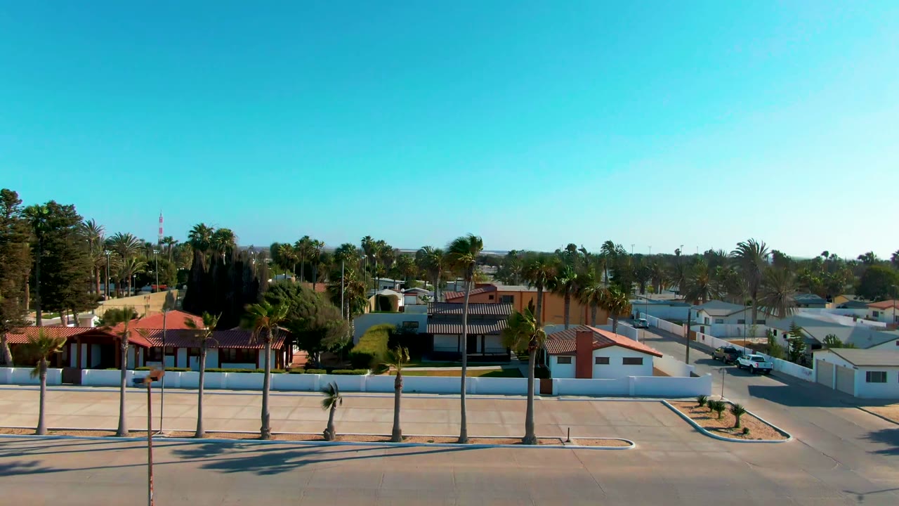 Early aerial frame over Guerrero Negro, Baja California Sur, showing red-roofed homes, white walls, palm trees, a street in the foreground, distant water on the horizon, and no visible people.