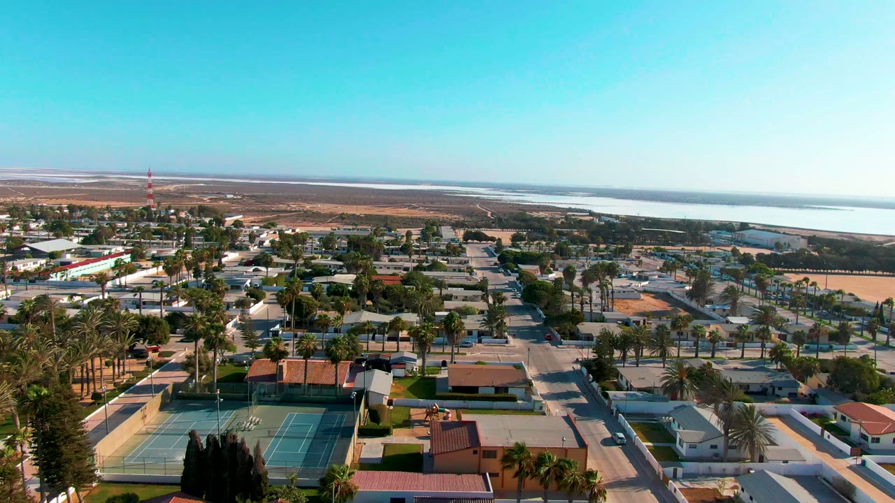 Opening aerial frame over Guerrero Negro, Baja California Sur, with houses, palm-lined roads, green tennis courts, dry land, distant water, and no visible people.