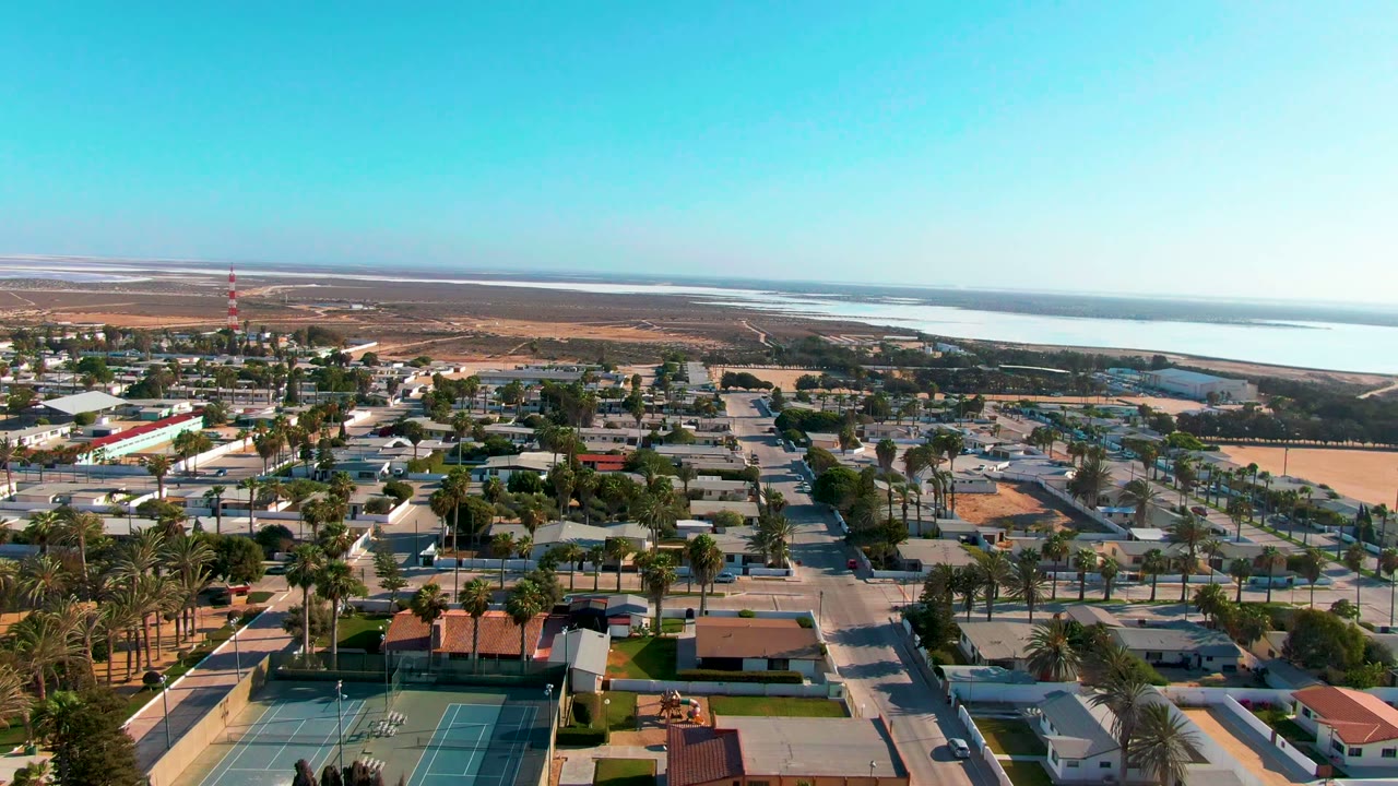 Mid-clip aerial frame over Guerrero Negro with rooflines and palms in the foreground, tennis courts near the lower left, open land behind town, distant water, and no visible people.