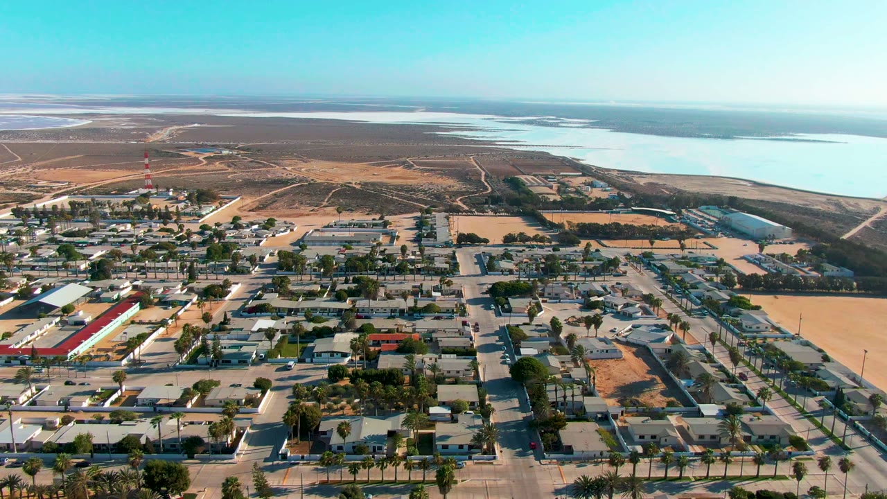 Mid-clip frame of Guerrero Negro with rooftops, straight roads, a red-and-white tower, brown open terrain, distant water, and no visible people.