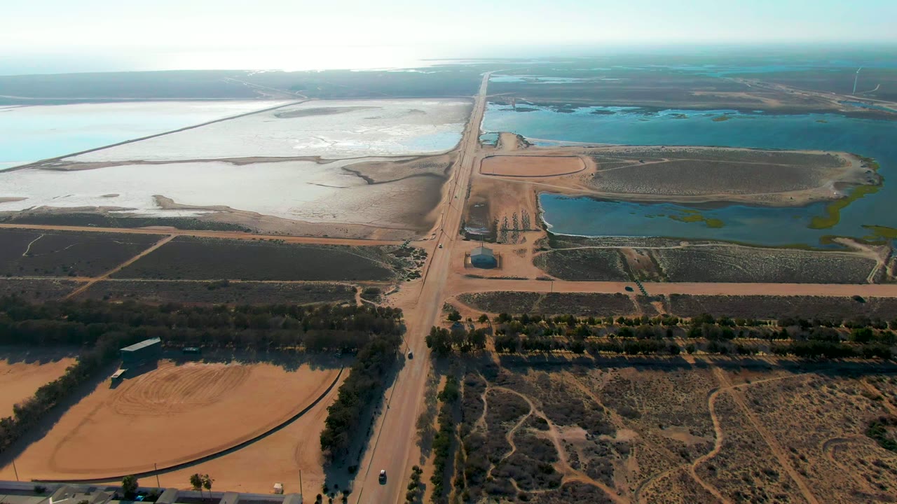 Aerial view over Guerrero Negro with a straight dirt road running upward between dry agricultural fields, blue reservoirs, pale water basins, and no visible people.
