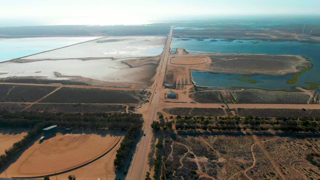 The aerial view over Guerrero Negro keeps the straight rural road near center, with tan earth, dark crop or scrub plots, blue water reservoirs, glare on pale basins, and no visible people.