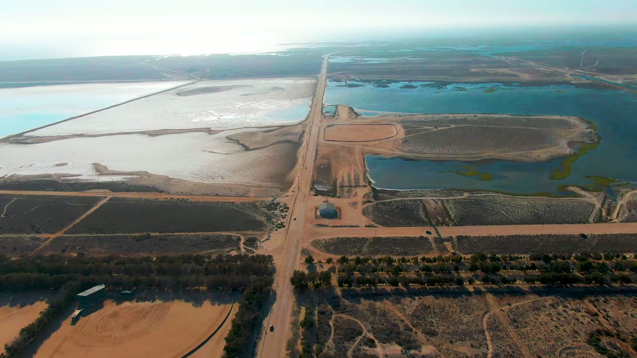 Aerial frame over Guerrero Negro, Baja California Sur, with a straight dirt road, dry fields, white flats, blue-green reservoirs, and no visible people.