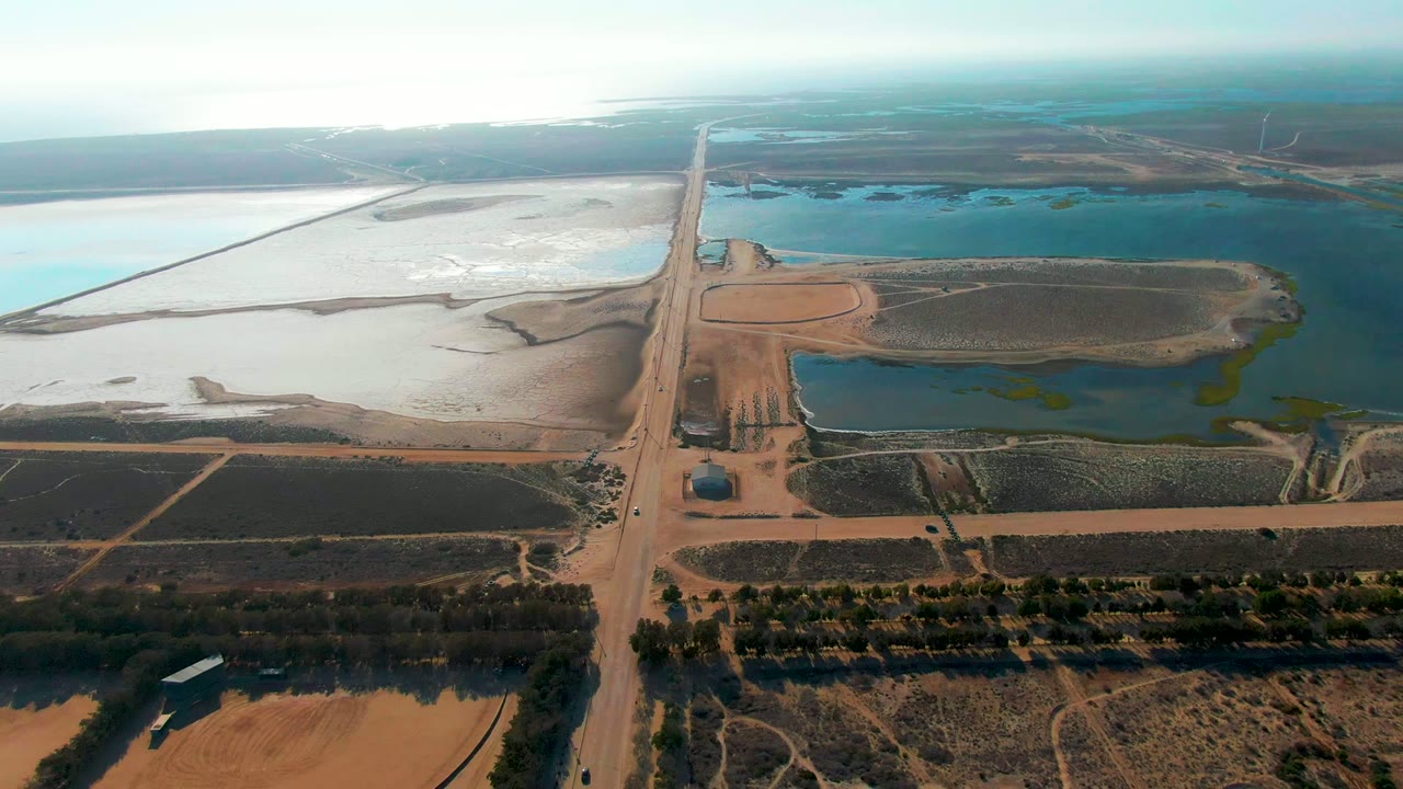 High oblique frame over Guerrero Negro, Baja California Sur, with the truck on the tan road, dark field blocks in the foreground, blue water to the right, white flats to the left, and no visible people.