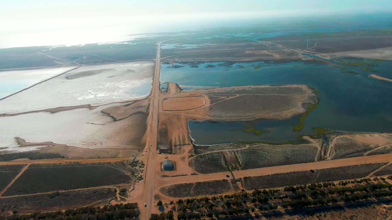 Aerial frame over Guerrero Negro, Baja California Sur, showing a straight dirt road between brown fields, blue-green ponds, pale flats, and no visible people.
