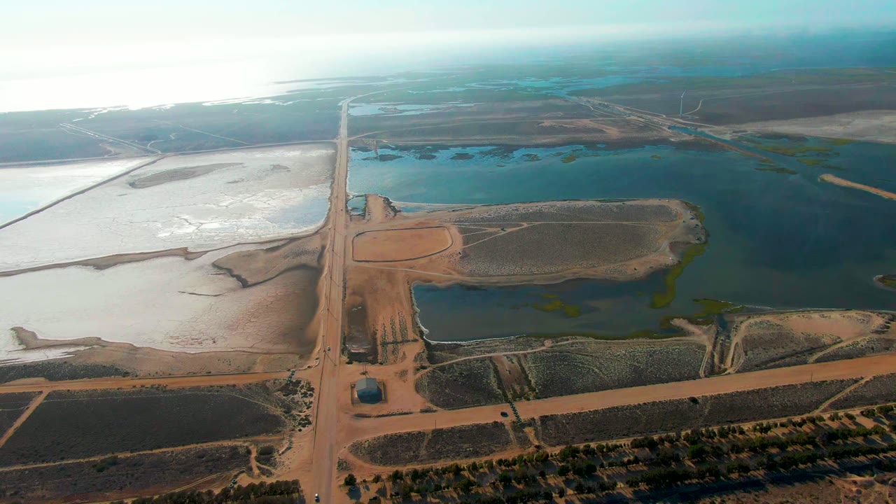 High drone-style frame over Guerrero Negro, Baja California Sur, with brown farm blocks in the foreground, water ponds beyond the road, bright flats to the left, and no visible people.