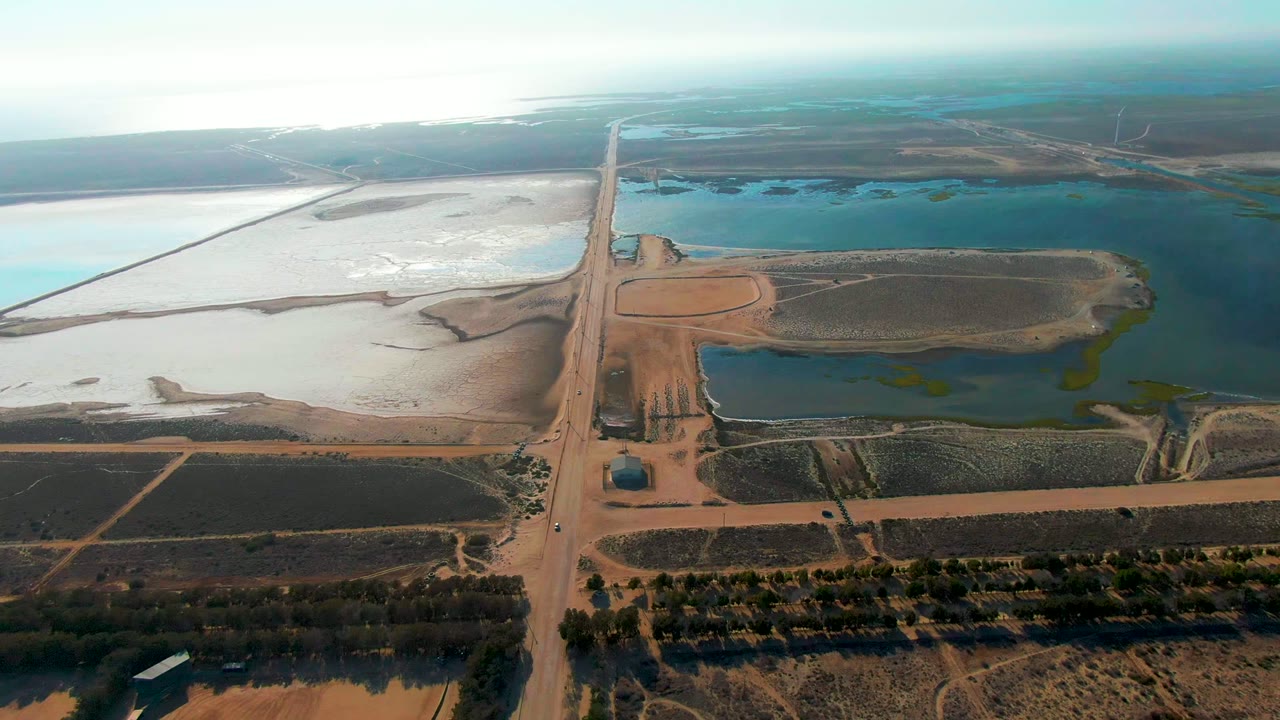High aerial view over Guerrero Negro, Baja California Sur, with a central dirt road crossing dry fields, blue-green ponds, pale flats, and no visible people.