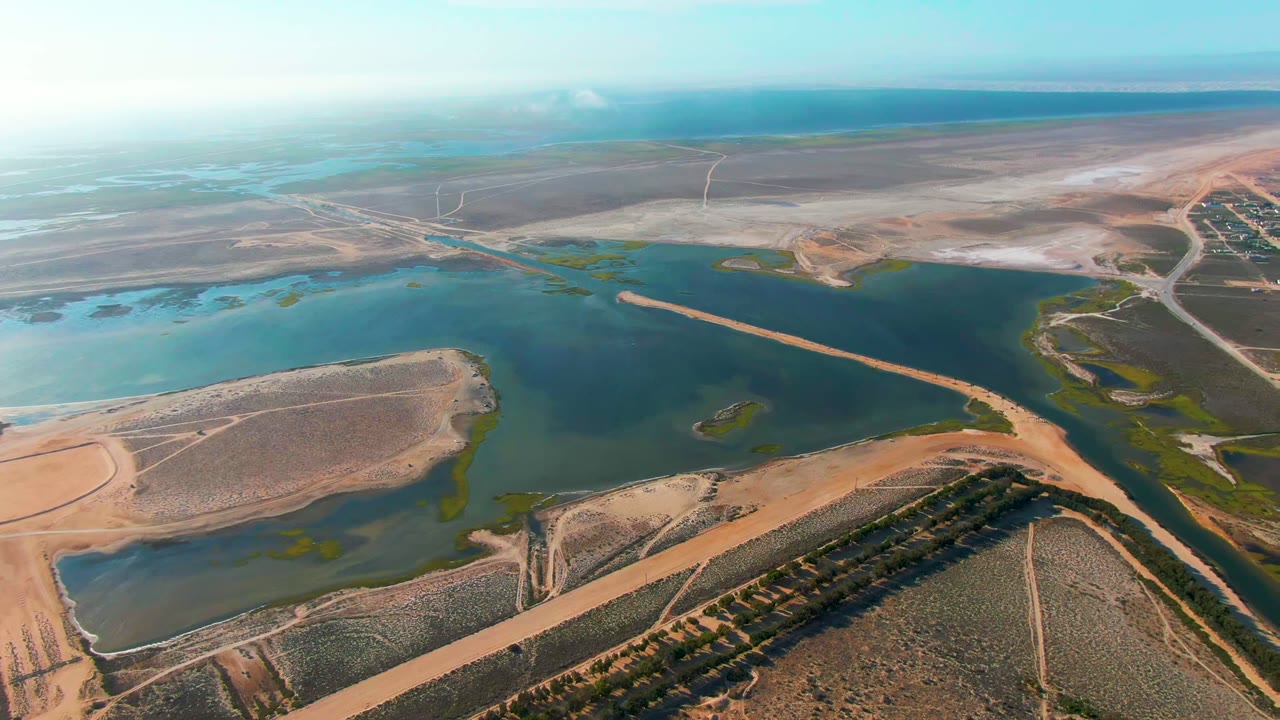 At 0:00, a high aerial view over Guerrero Negro shows blue-green reservoir water, dry brown fields, dirt roads, and a distant flat horizon with no visible people.