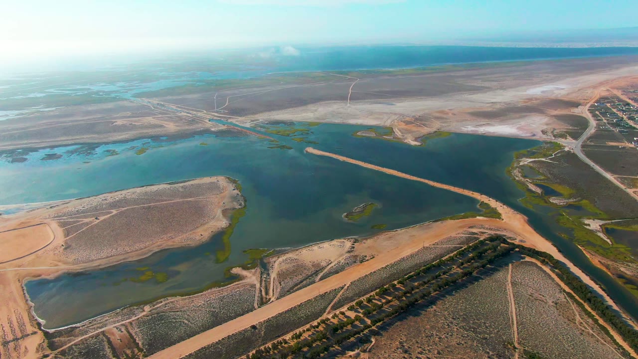 Drone view of reservoir, embankment, farmland, and coastal-looking water beyond.