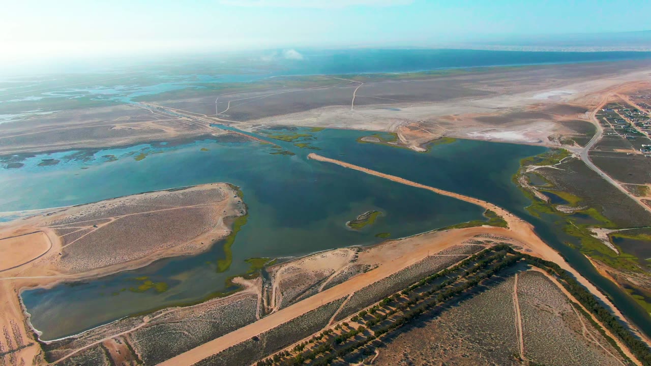 At 0:02, the drone view frames the reservoir center with curved shoreline vegetation, tan access roads, dry fields, and broad blue sky over Guerrero Negro, with no visible people.