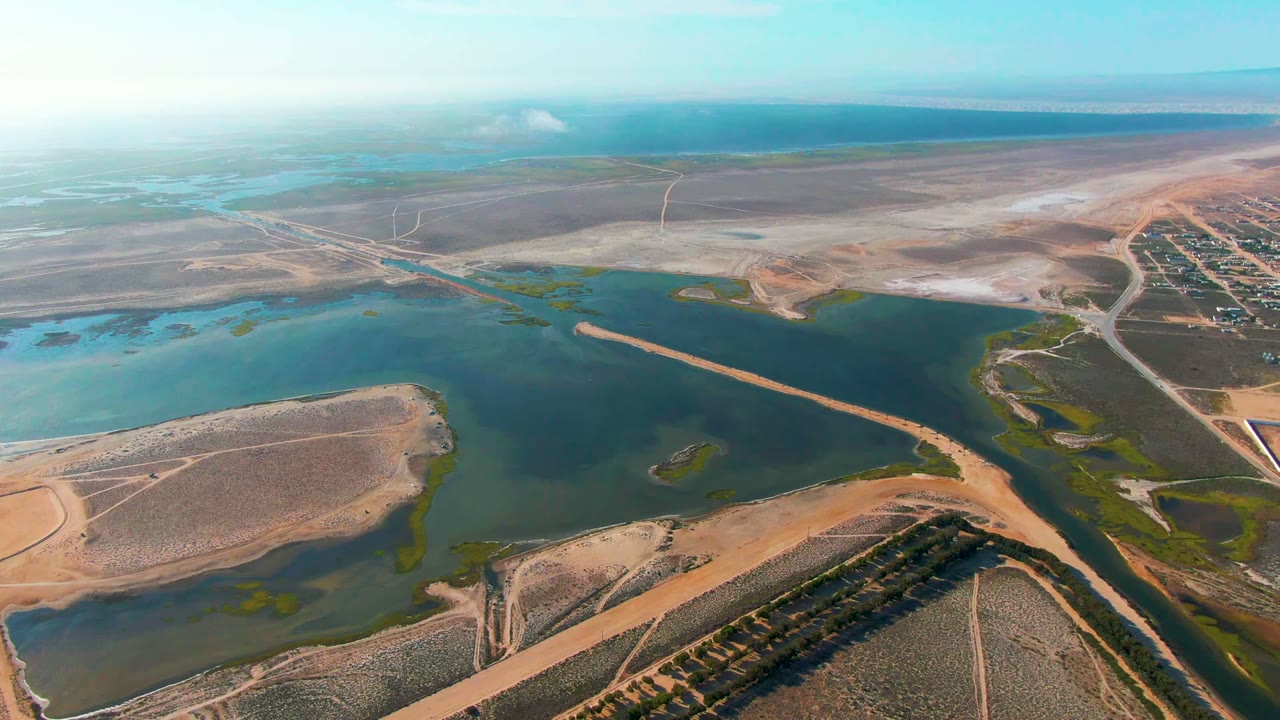 Aerial view over a green-blue reservoir in Guerrero Negro, Baja California Sur, with sandy roads, dry flats, wetlands, distant horizon haze, and no visible people.