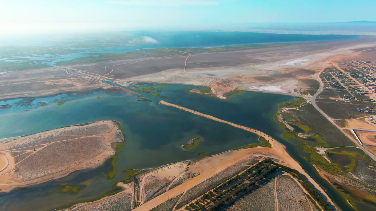 Aerial frame over Guerrero Negro, Baja California Sur, showing blue-green water bordered by tan dry land, roads, a developed grid at right, distant water on the horizon, and no visible people.
