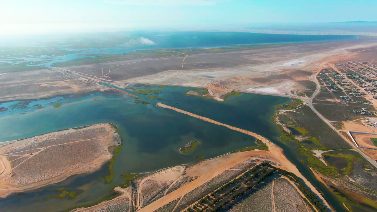 A Guerrero Negro aerial frame shows a road or causeway along the blue-green water, tan shorelines, green wetland patches, a developed grid to the right, and no visible people.