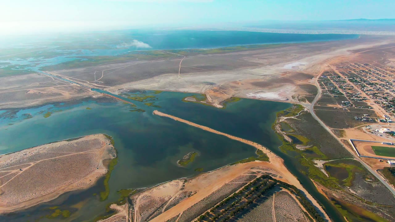 Opening aerial frame over Guerrero Negro wetlands and arid terrain, showing blue-green water, tan sandbars, roads, a small settlement to the right, haze on the horizon, and no visible people.