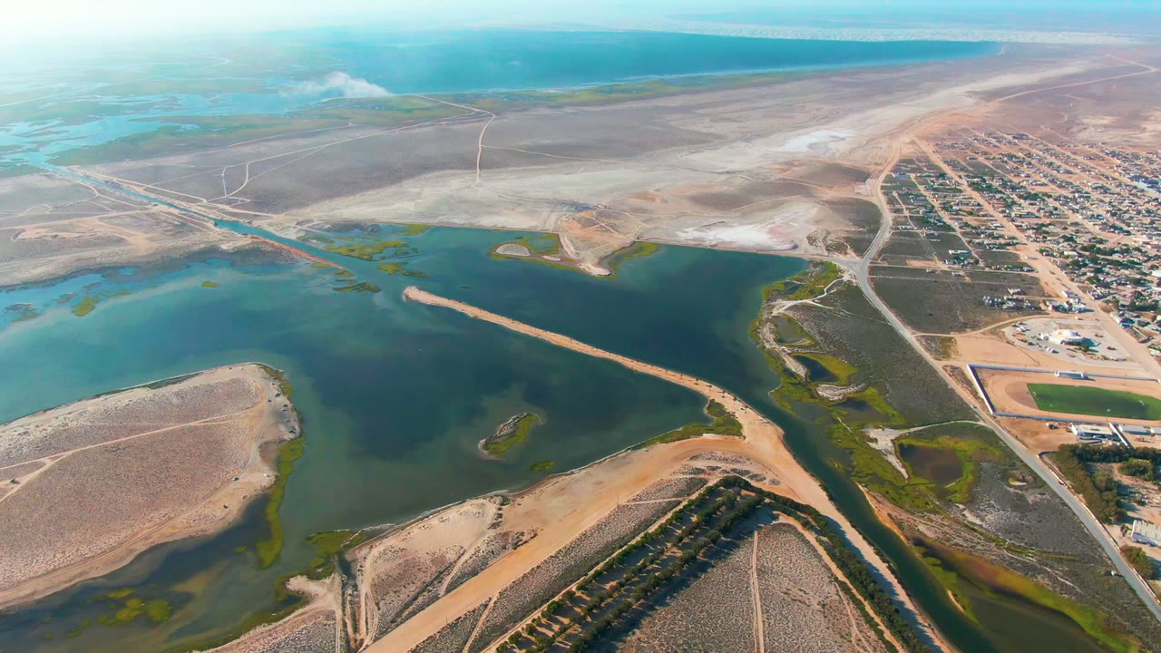 Opening aerial frame over Guerrero Negro, Baja California Sur, Mexico, with calm green-blue water, tan dry land, a town grid at right, and a small smoke or steam plume near the upper left, no visible people.